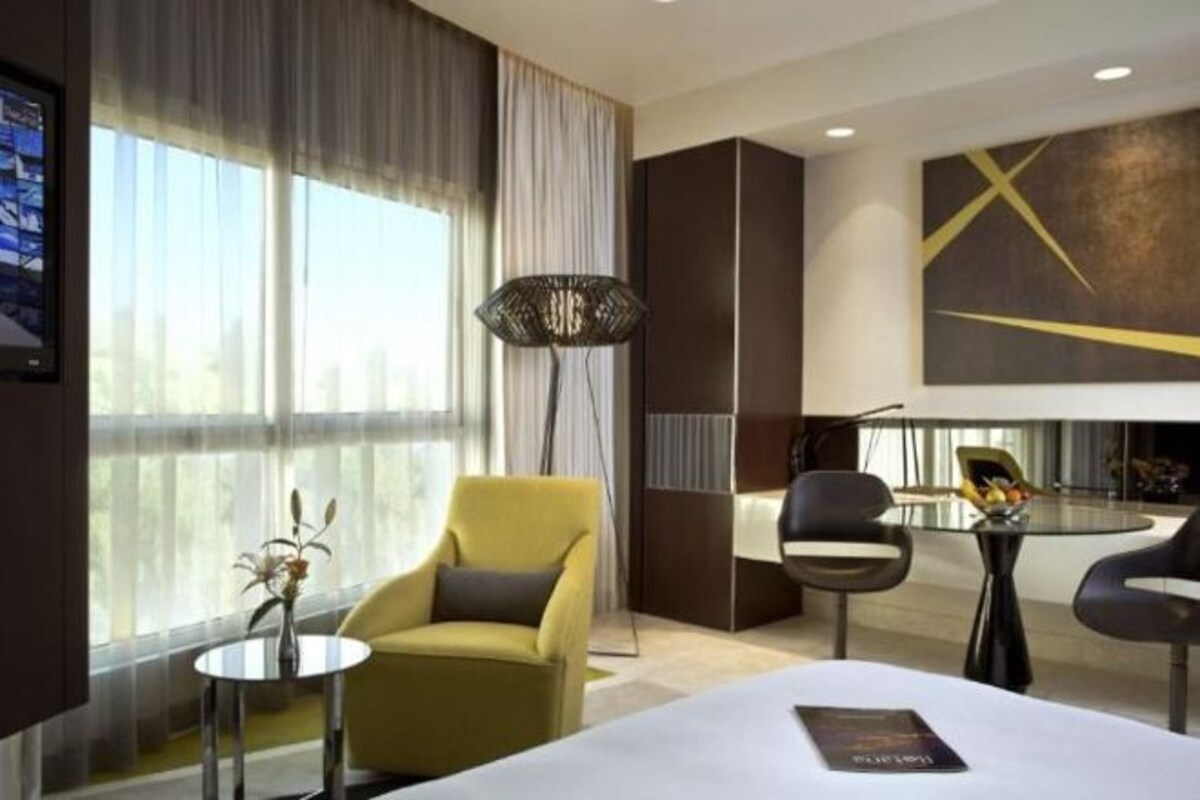 A modern hotel room with a large bed, a desk, a chair, and a view of the city through large windows.