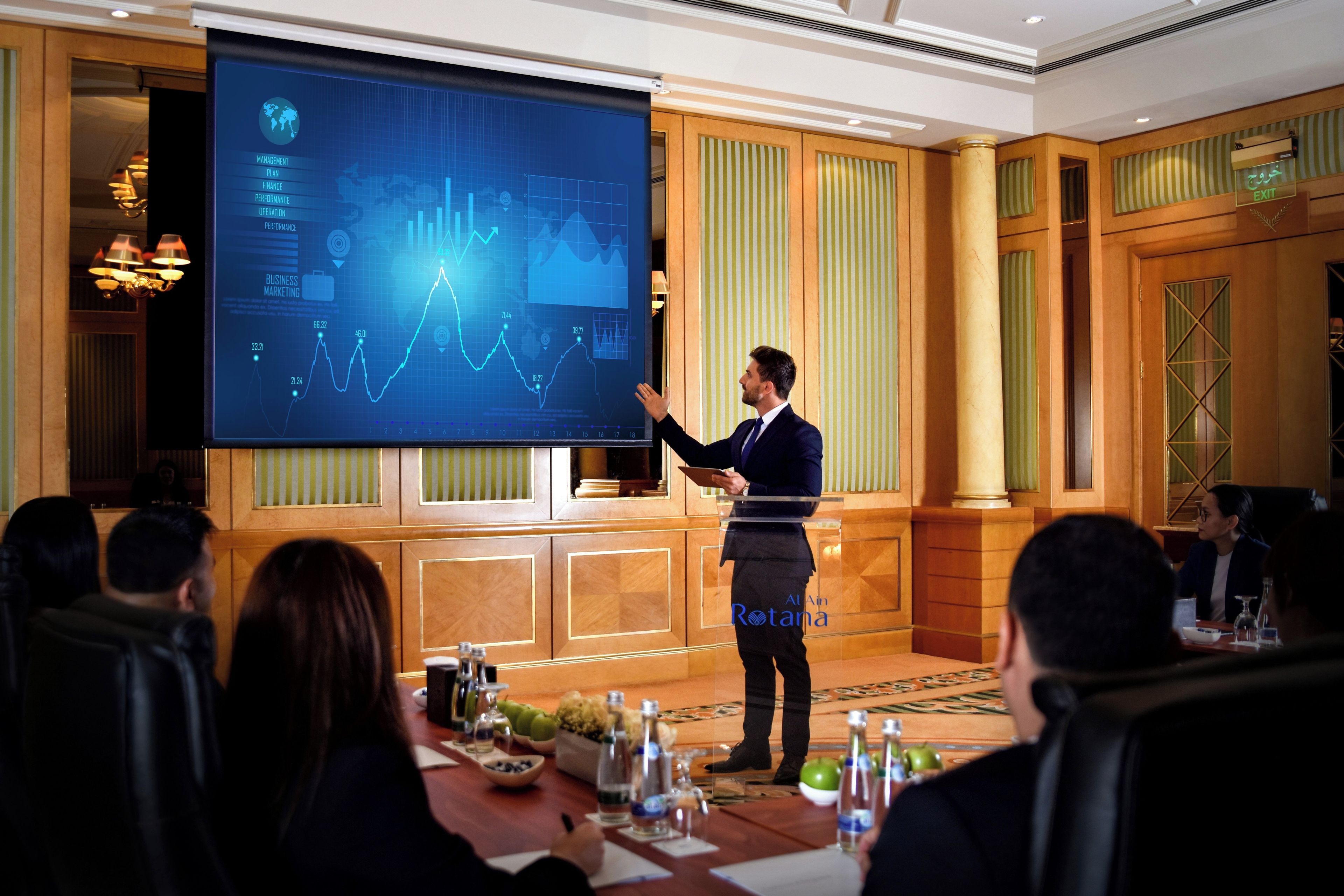 A conference room with a presentation screen displaying graphs and charts, a podium, and attendees seated at a table with water bottles and notepads.
