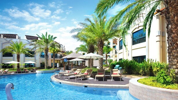 A swimming pool with lounge chairs and umbrellas, surrounded by palm trees and a building with balconies. - Al Ain Rotana (Al Ain)