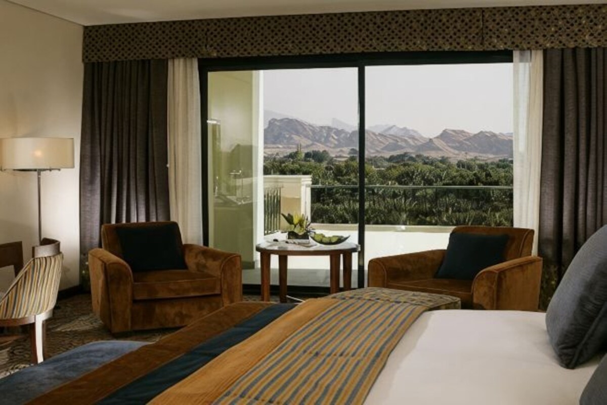 A hotel room with a large bed, two armchairs, a small table with a fruit bowl, and a view of mountains through a sliding door.