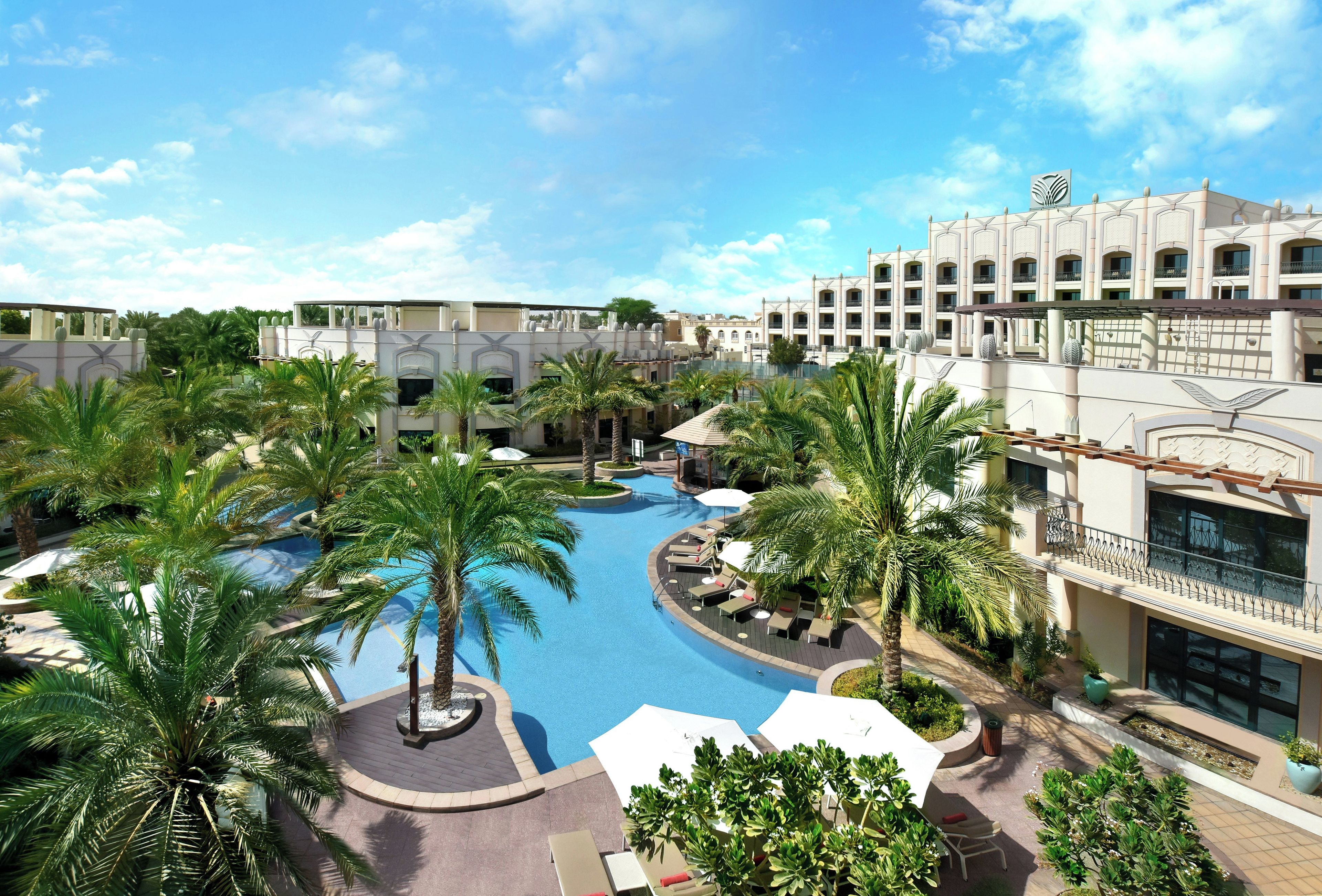 Resort com piscina central, palmeiras e um edifício de vários andares com varandas.