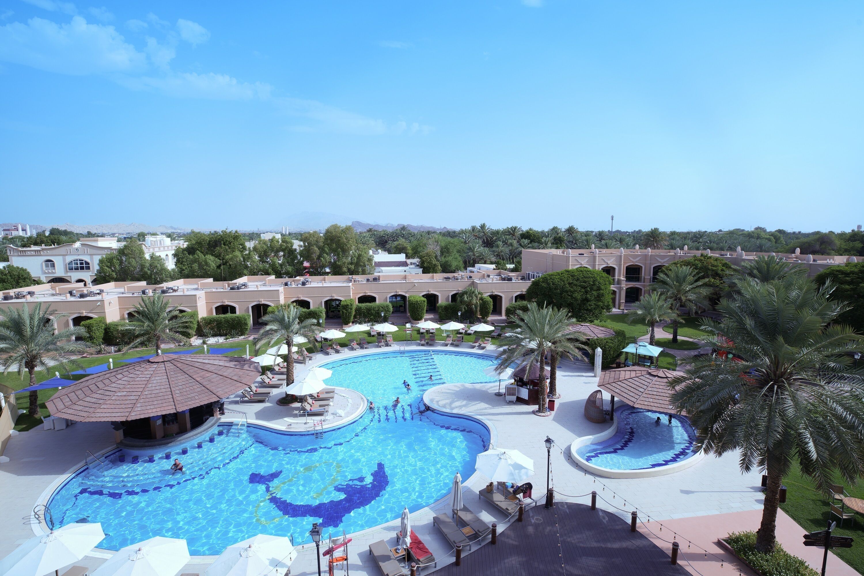 A large outdoor swimming pool with multiple sections, surrounded by palm trees and lounge areas.