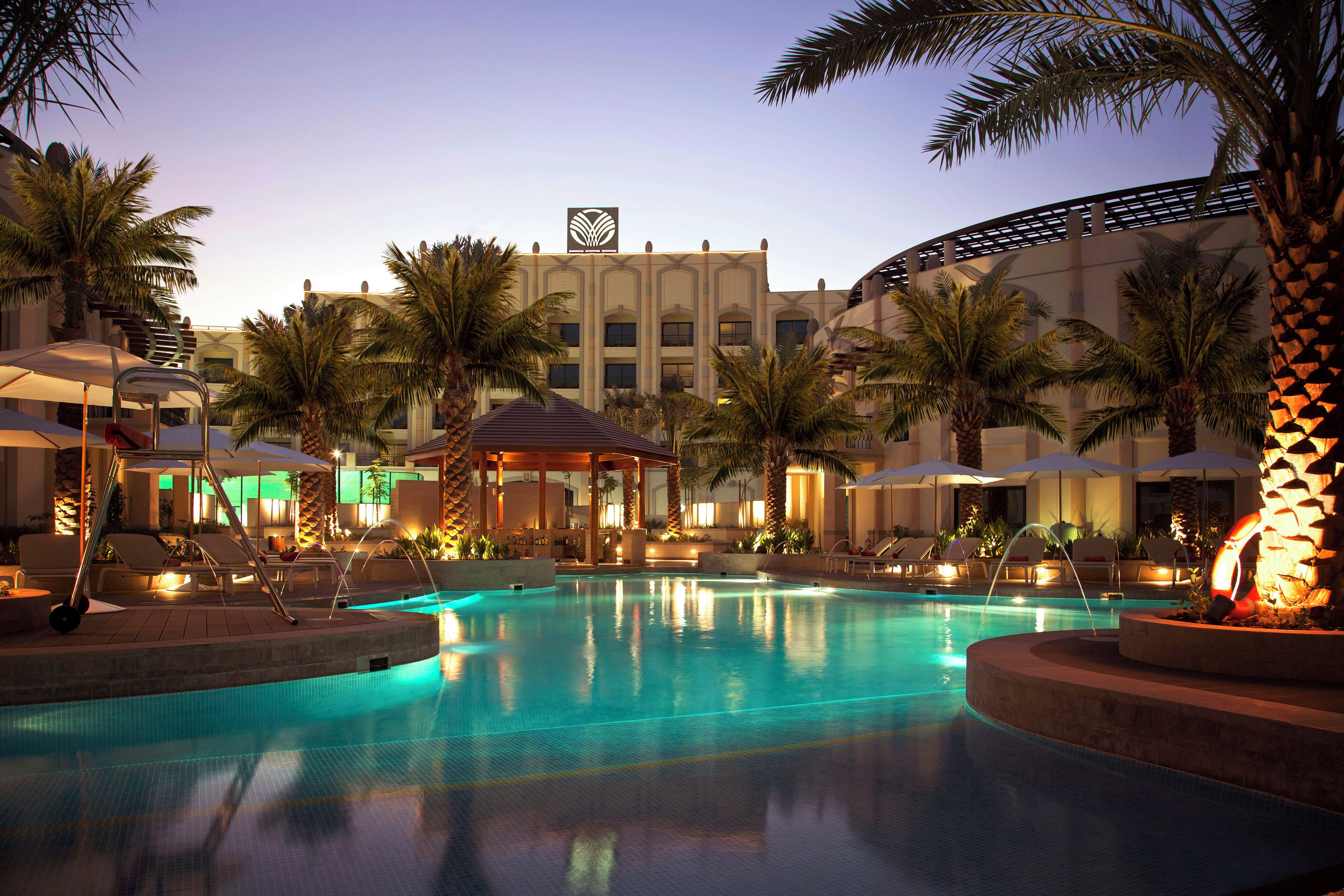 Área de piscina de hotel com espreguiçadeiras, palmeiras e um edifício com um logotipo característico.