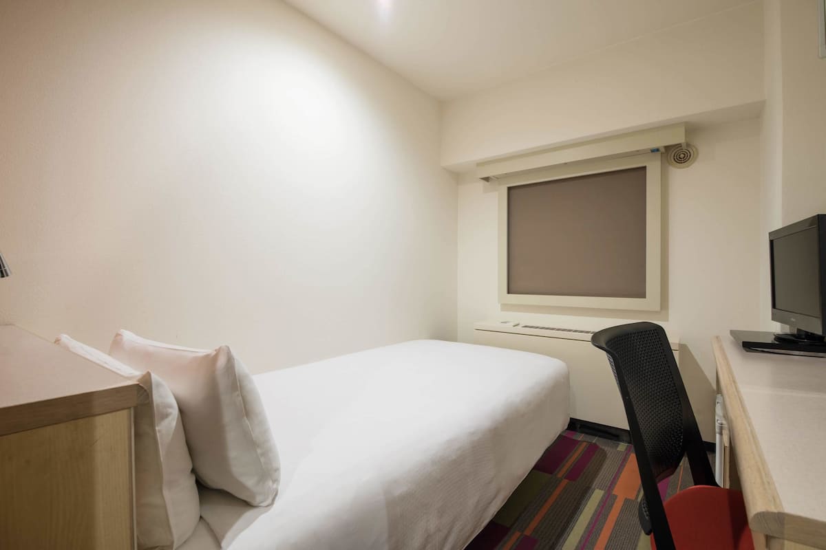 A hotel room with a single bed, a desk with a computer, and a window with a blind.