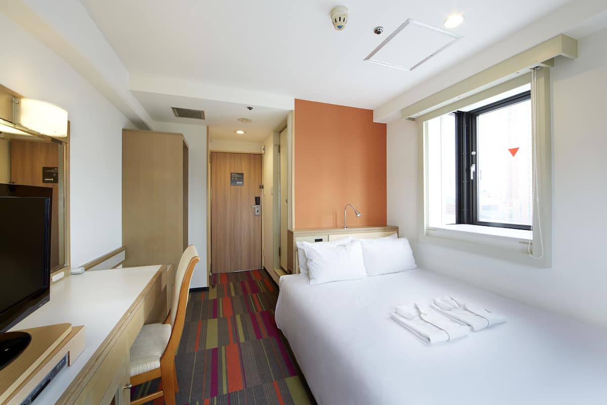 A hotel room with a bed, a desk with a chair, a television, and a window with curtains.