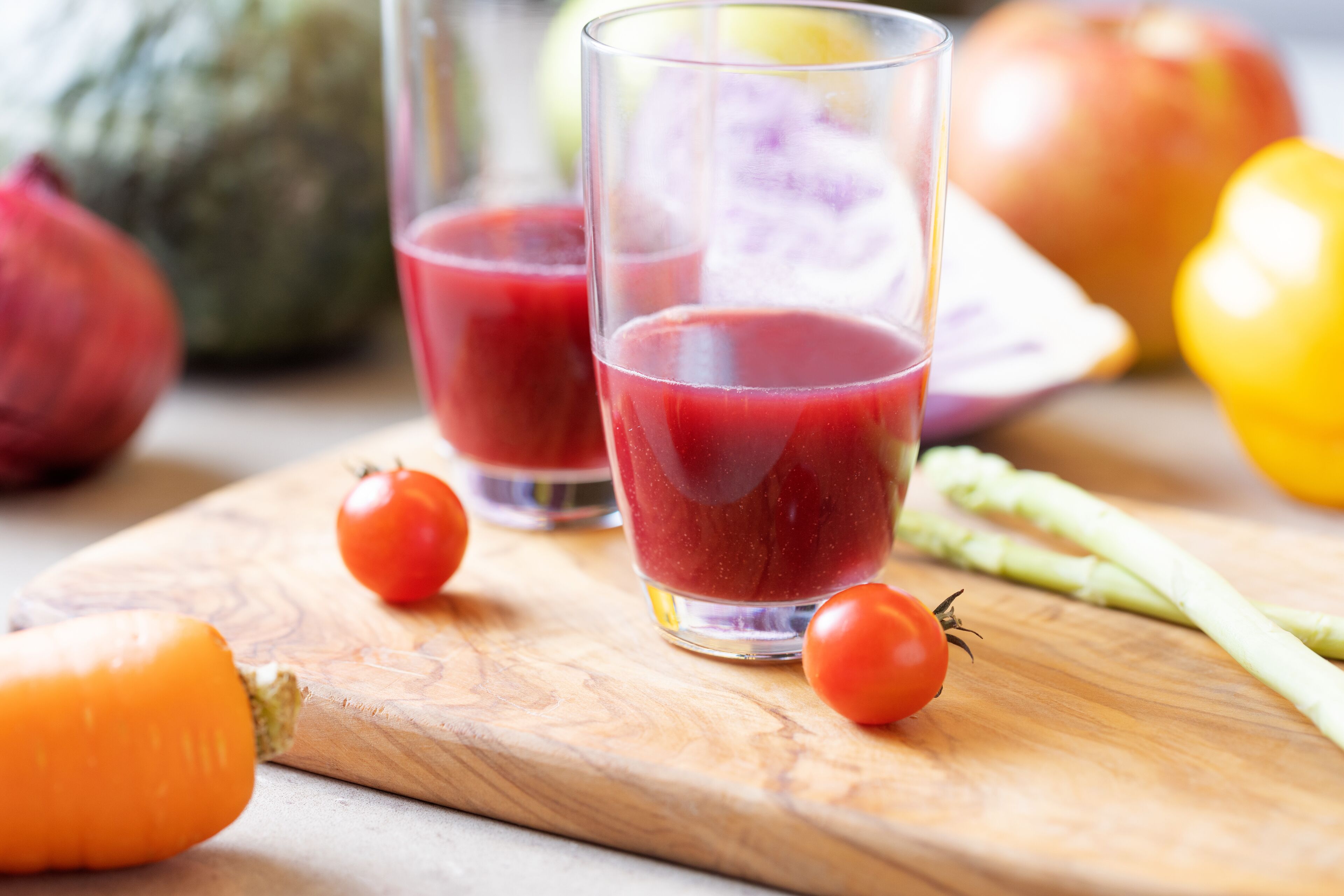 Dos vasos de jugo rojo sobre una tabla de madera con verduras a su alrededor.