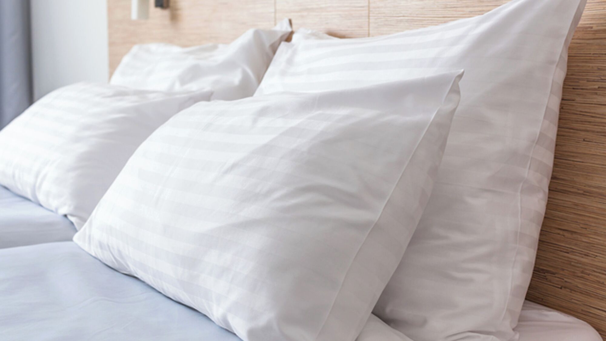 A neatly made bed with white bedding and pillows, a wooden headboard, and a light-colored wall.