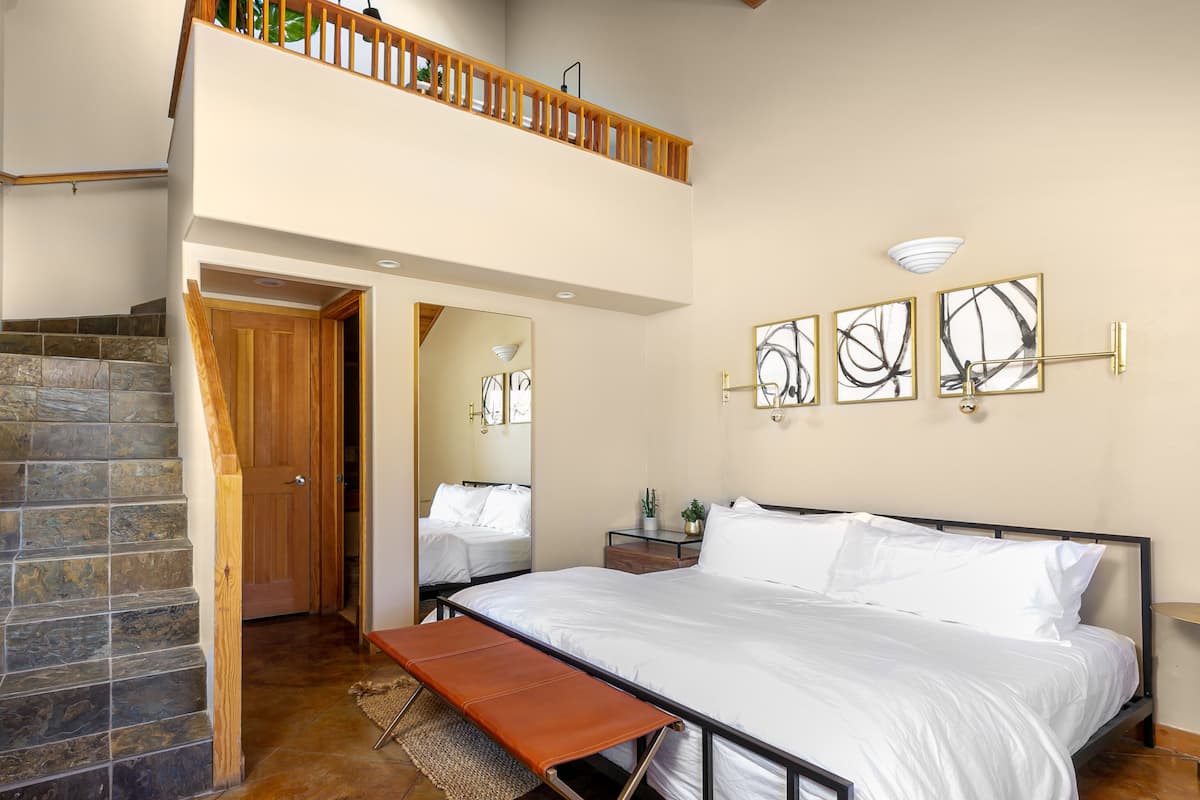 A bedroom with a large bed, a wooden bench, a stone staircase, and a balcony with a railing.