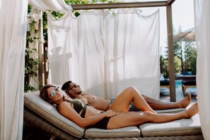 Un couple se détend sur un fauteuil confortable installé à l’extérieur, sous une pergola, avec une piscine en arrière-plan.