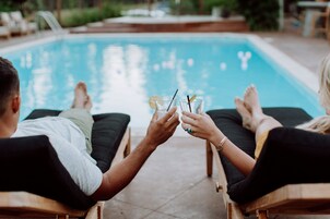 Dos personas sentadas junto a una piscina, con bebidas, sillas de descanso y vista despejada a la piscina.