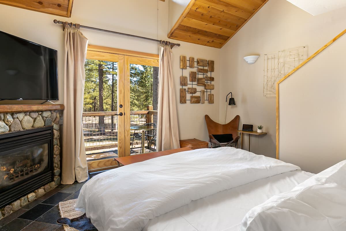 A bedroom with a stone fireplace, a bed, a TV, a desk, and a view of the outdoors.