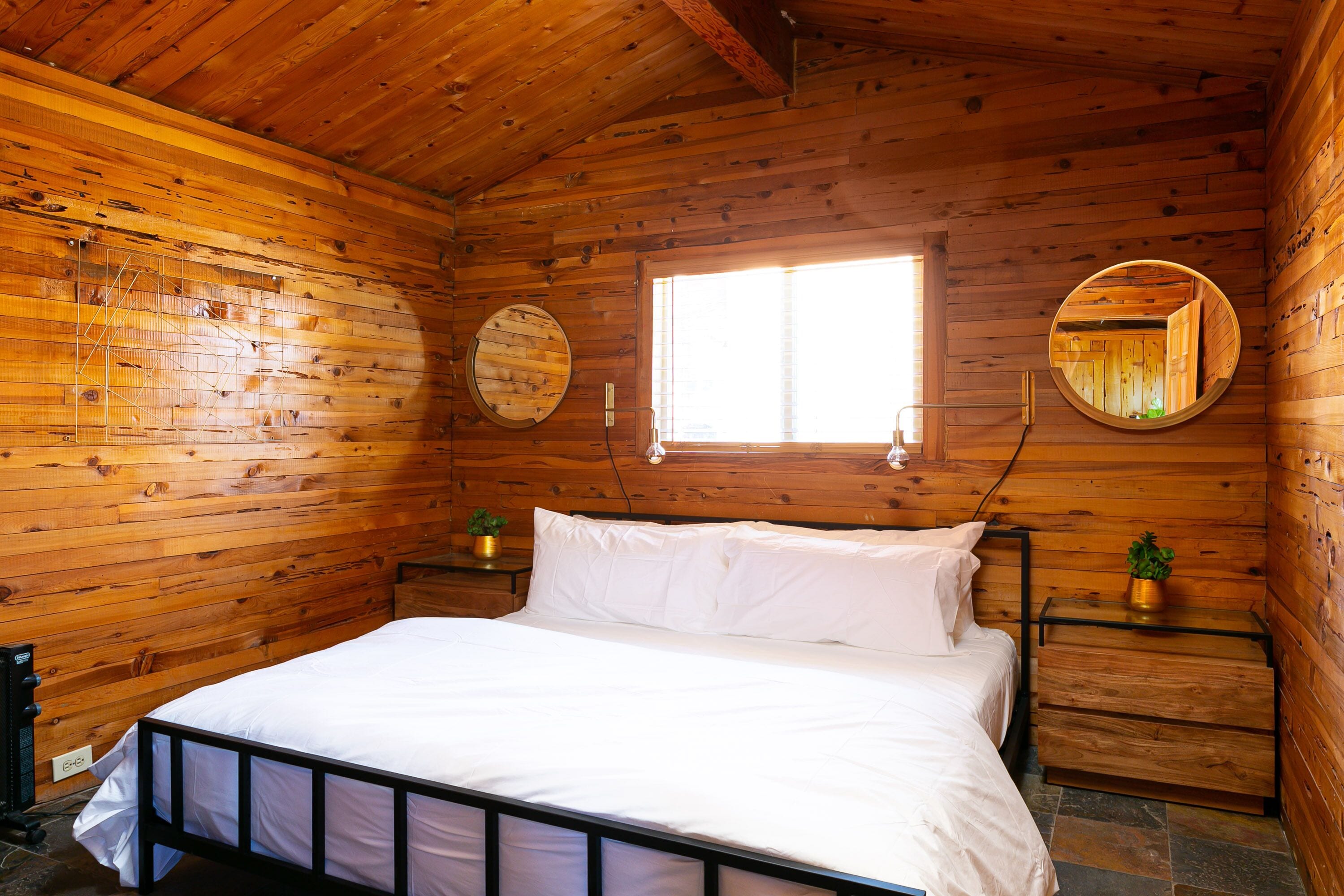 A wooden cabin bedroom with a bed, two bedside tables, a window, and two wall-mounted mirrors.