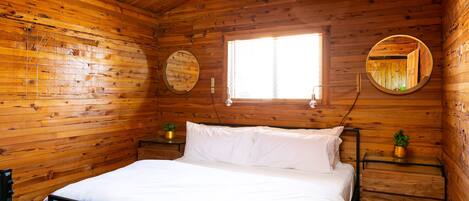 A wooden cabin bedroom with a bed, two bedside tables, a window, and two wall-mounted mirrors.