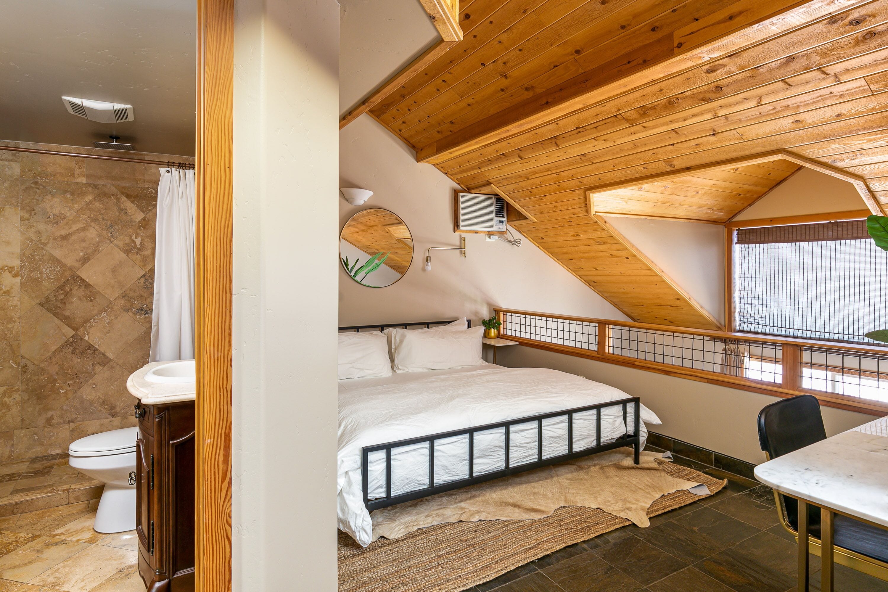 Un dormitorio con techo de madera, una cama, un escritorio pequeño y una ventana con persianas.