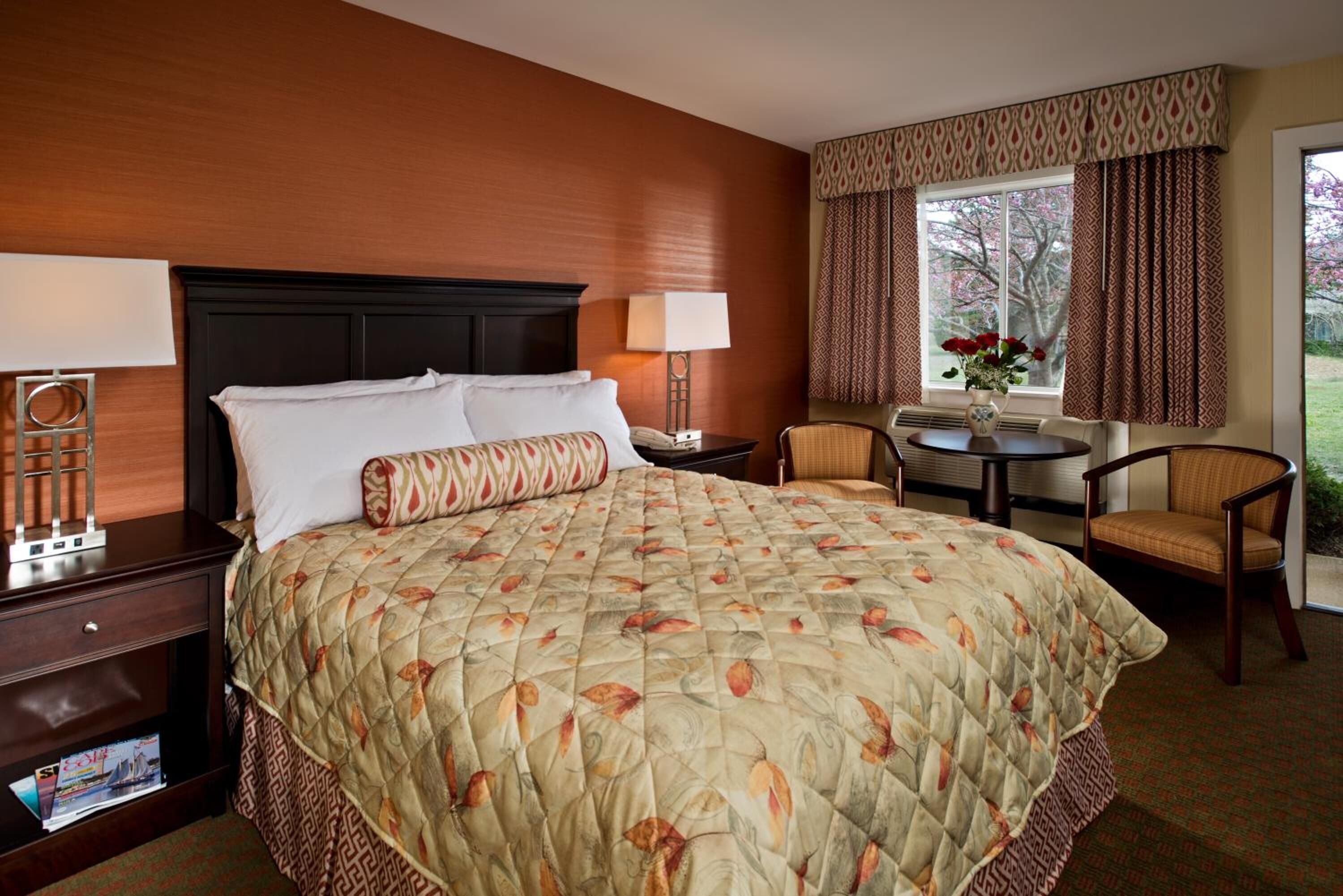 Une chambre d’hôtel avec un grand lit, une table de chevet, une chaise, une petite table et une fenêtre avec des rideaux.