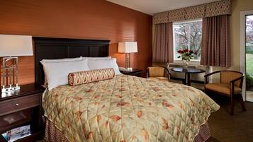Une chambre d’hôtel avec un grand lit, une table de chevet, une chaise, une petite table et une fenêtre avec des rideaux.