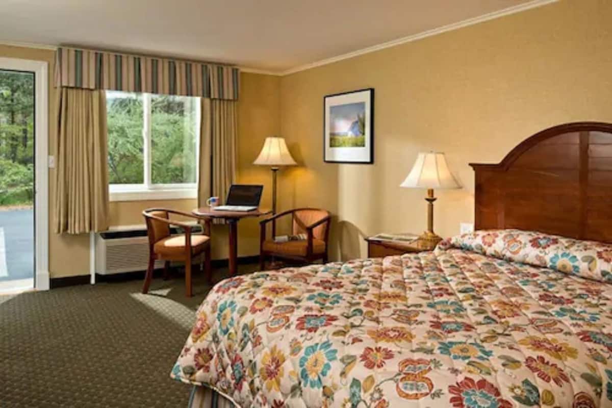 A hotel room with a bed, two chairs, a desk, a laptop, a lamp, and a window with curtains.