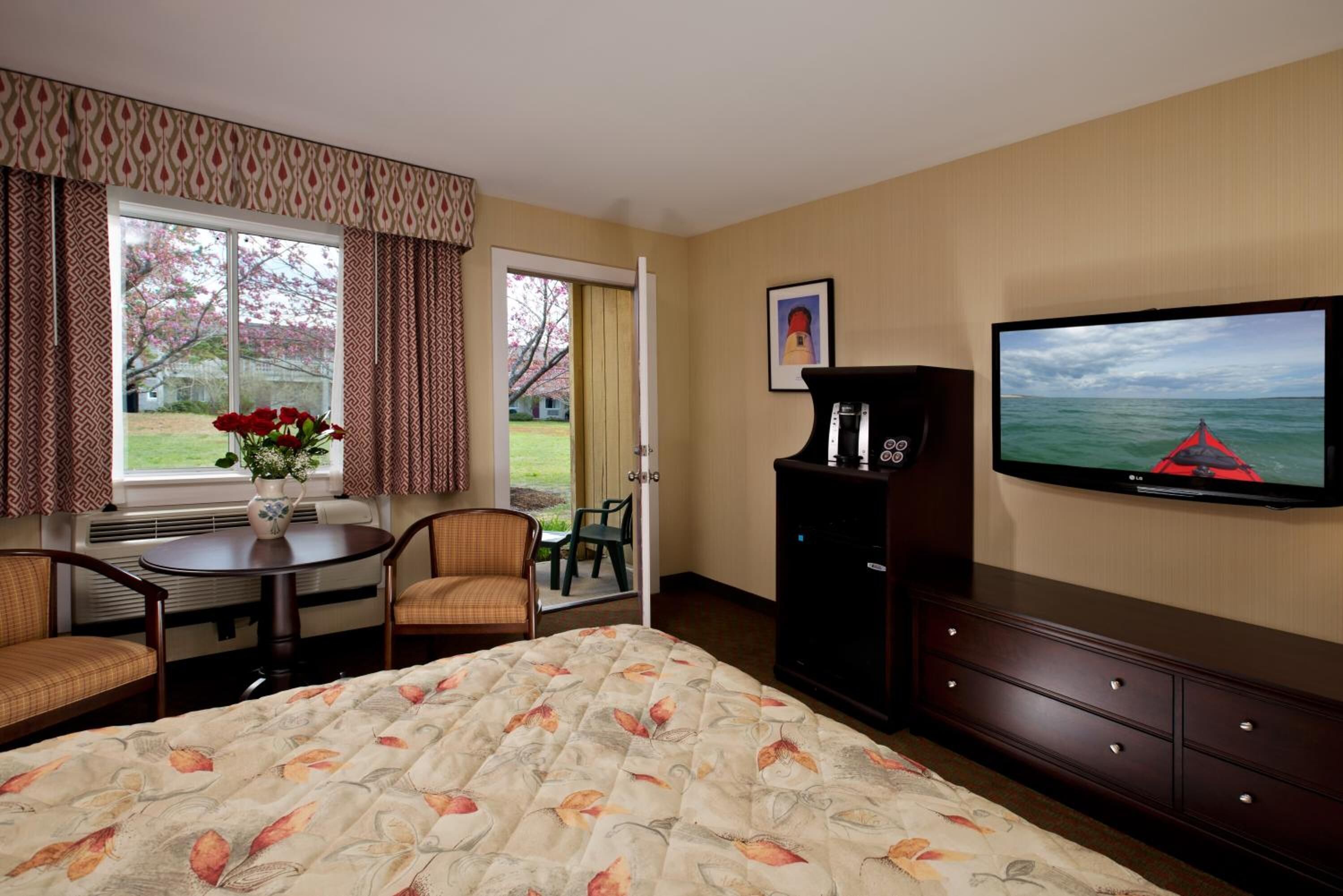 A hotel room with a bed, a TV, a desk with a chair, a small table with a vase of flowers, and a view of the outdoors through a window.