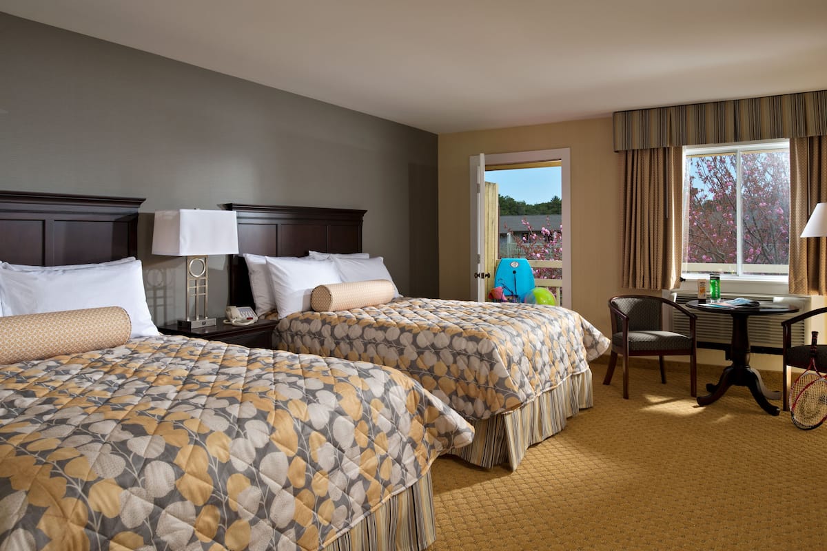 A hotel room with two beds, a desk, a chair, and a window with a view of trees.