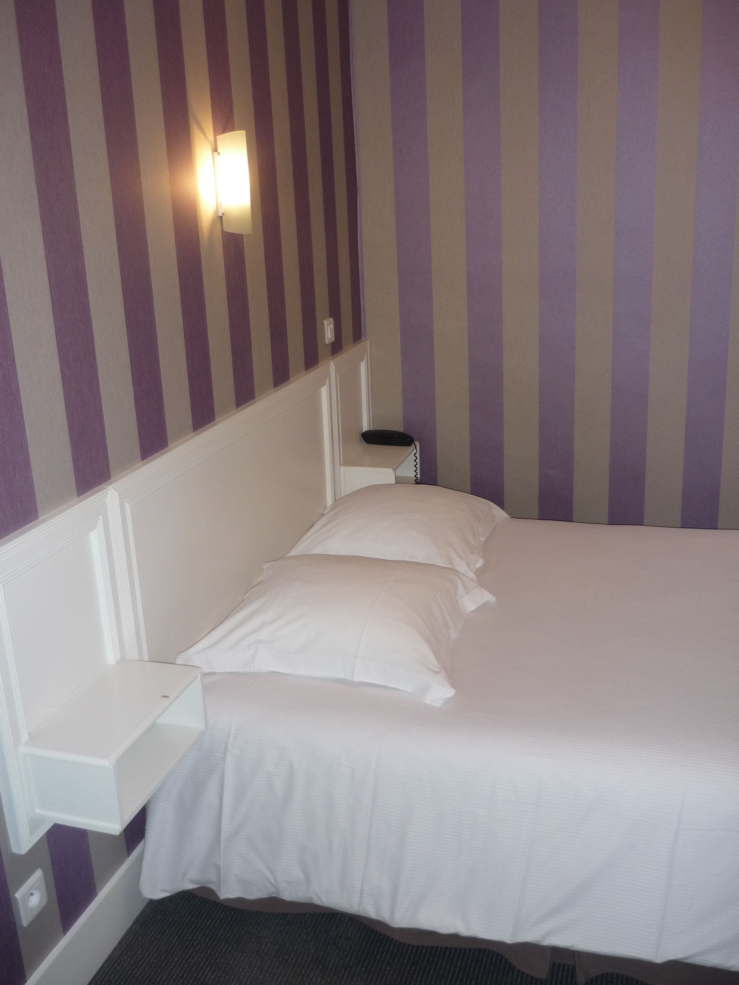 A hotel room with a bed, a nightstand, and a wall-mounted lamp.