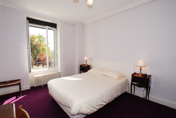 A neatly arranged bedroom with a large bed, two bedside tables with lamps, a radiator, a bench, and a window with a view of trees.