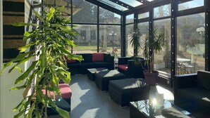 A conservatory with glass walls, furnished with sofas and a table, and surrounded by potted plants.