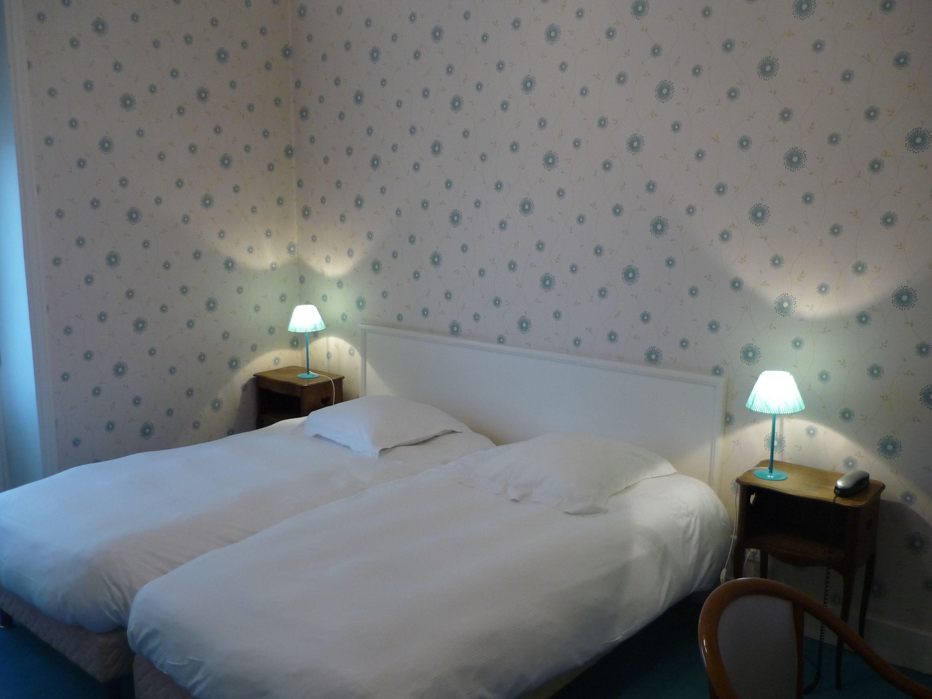 A neatly made bed with white linens in a room with floral wallpaper. There are two bedside tables, each with a lamp. A wooden chair is positioned near the bed.
