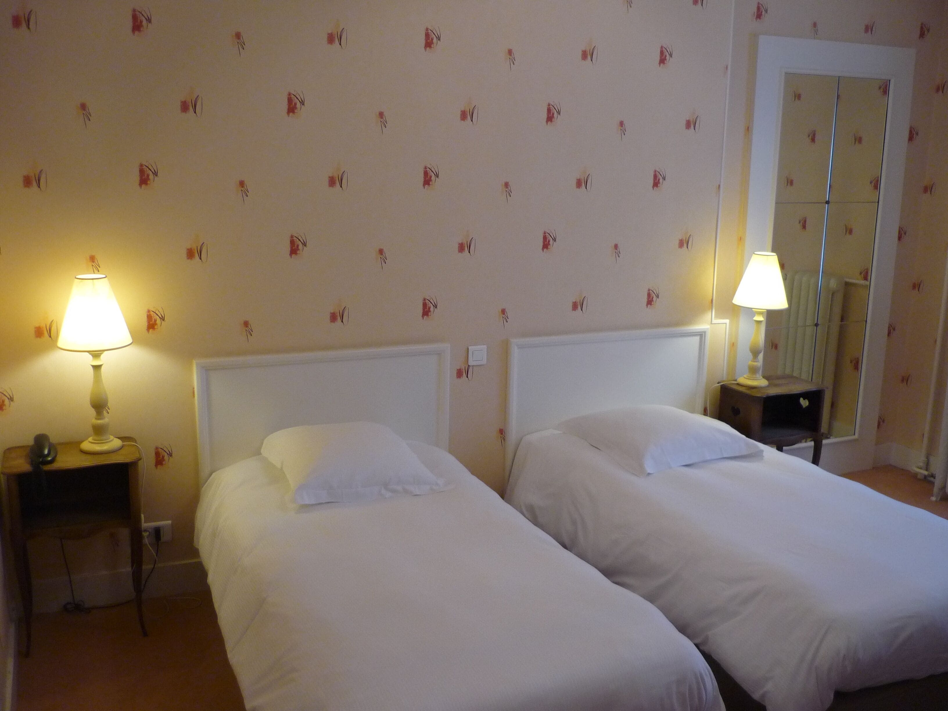 A hotel room with two single beds, each with a white headboard and pillow. There are two bedside tables, one on each side of the beds, each with a lamp. The walls have a floral wallpaper pattern.