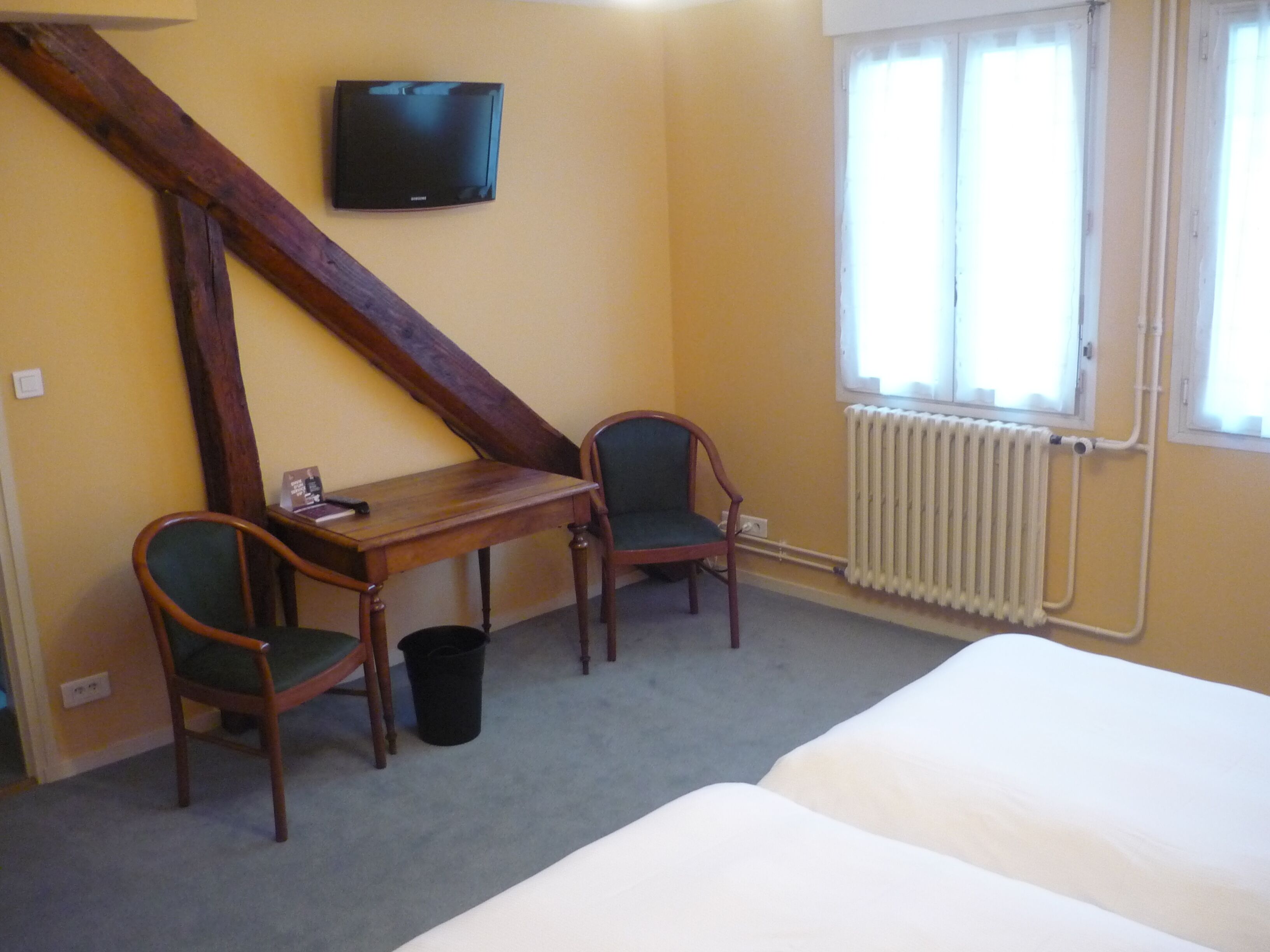Een kamer met een bed, twee stoelen, een klein tafeltje, een televisie aan de muur en een raam met een radiator eronder.