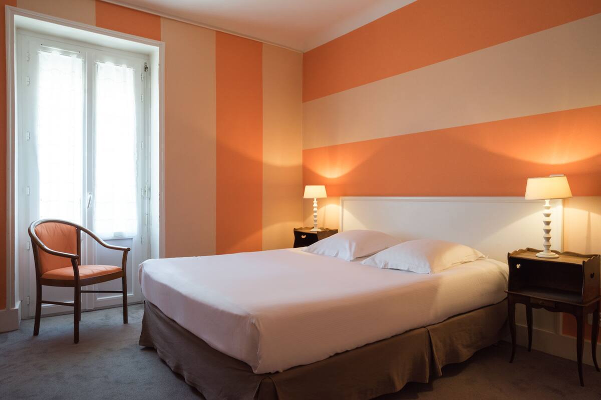 A hotel room with a bed, a chair, two bedside tables with lamps, and a window with curtains.