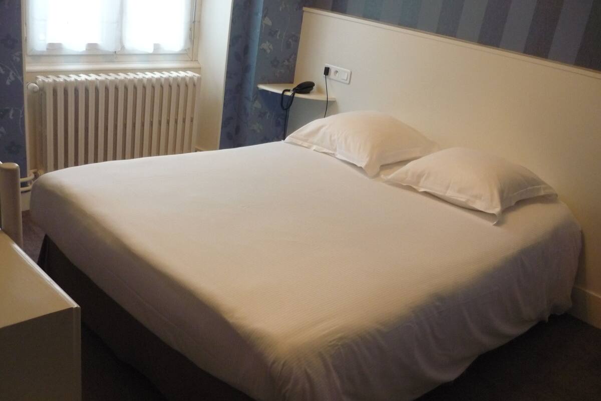 A neatly made bed with white linens in a room with a radiator, a power outlet, and a blue-striped wall.