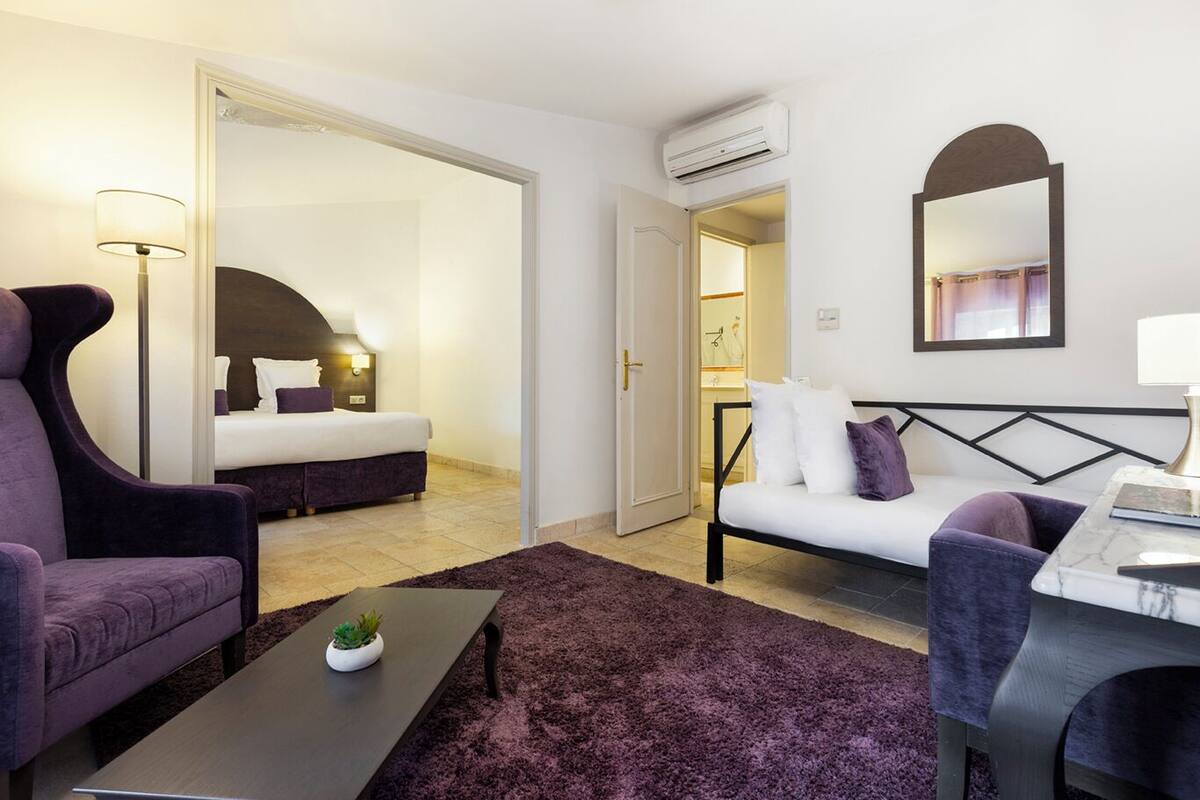 A hotel room with a bed, a sofa, a chair, a small table with a plant, and a mirror on the wall.