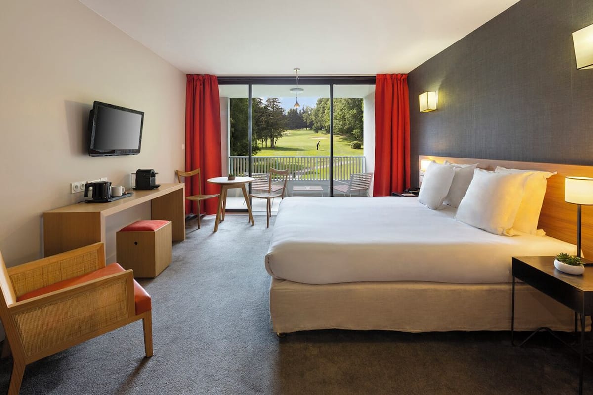 A hotel room with a large bed, a desk with a television, a chair, a small table, and a view of a golf course through the window.