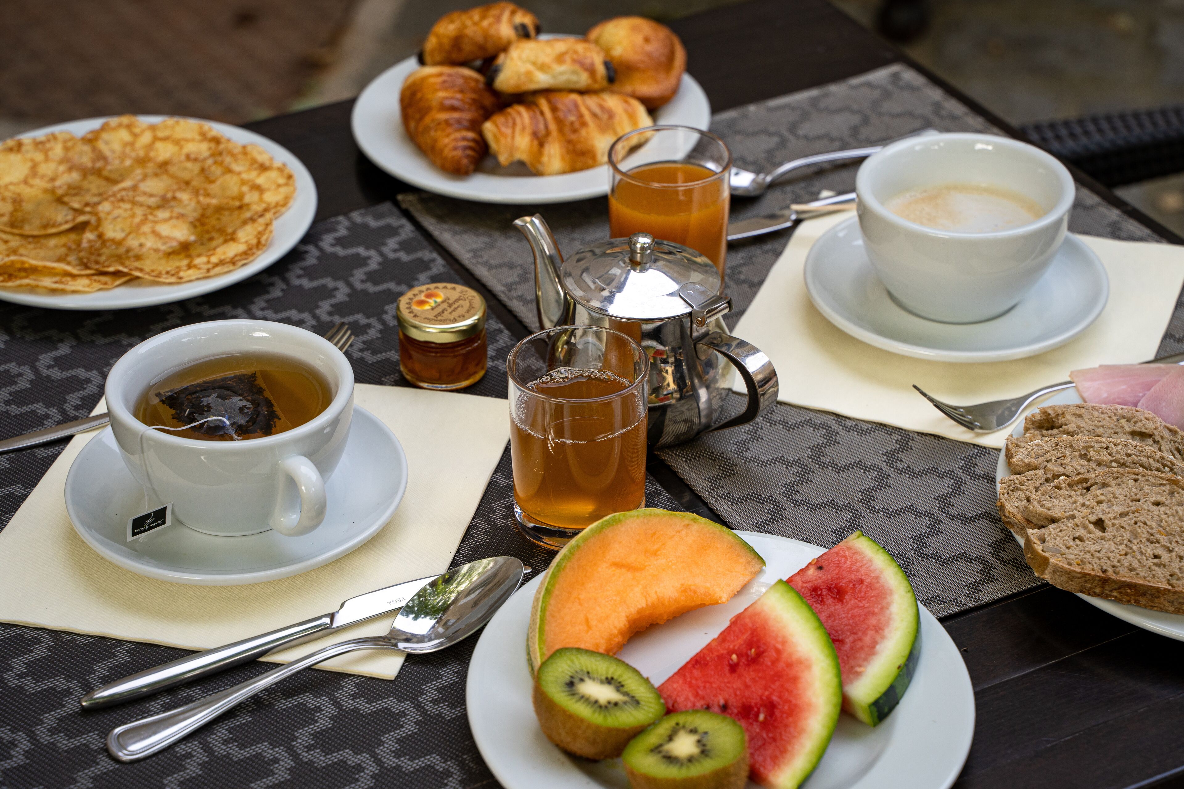A breakfast setting with croissants, a plate of sliced watermelon, kiwi, and bread, along with cups of tea and coffee.