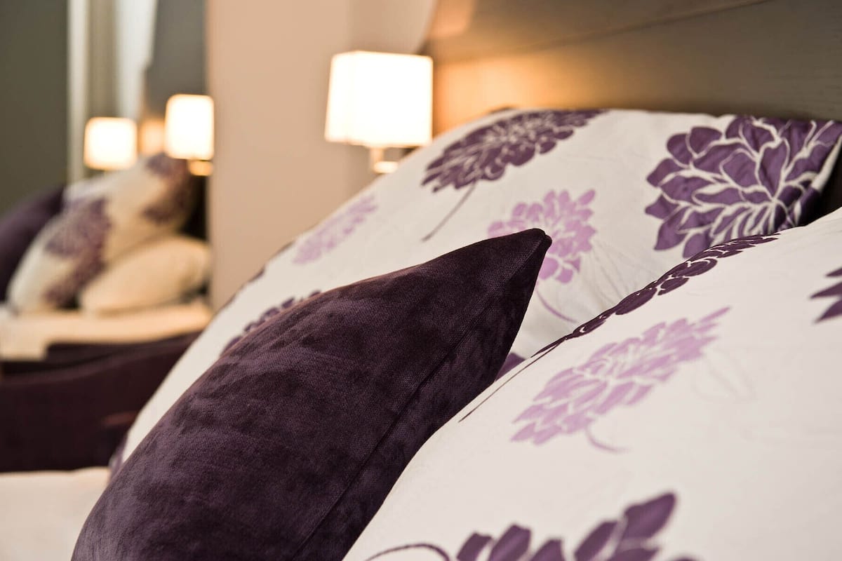 A close-up of a bed with purple floral pillows and a dark purple throw.