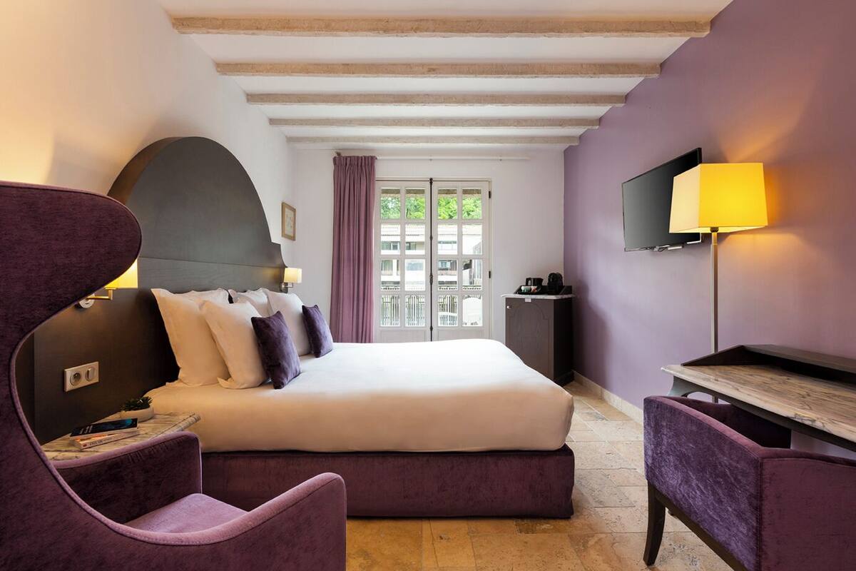 A bedroom with a bed, a desk, a chair, a TV, and a window with a view of buildings.