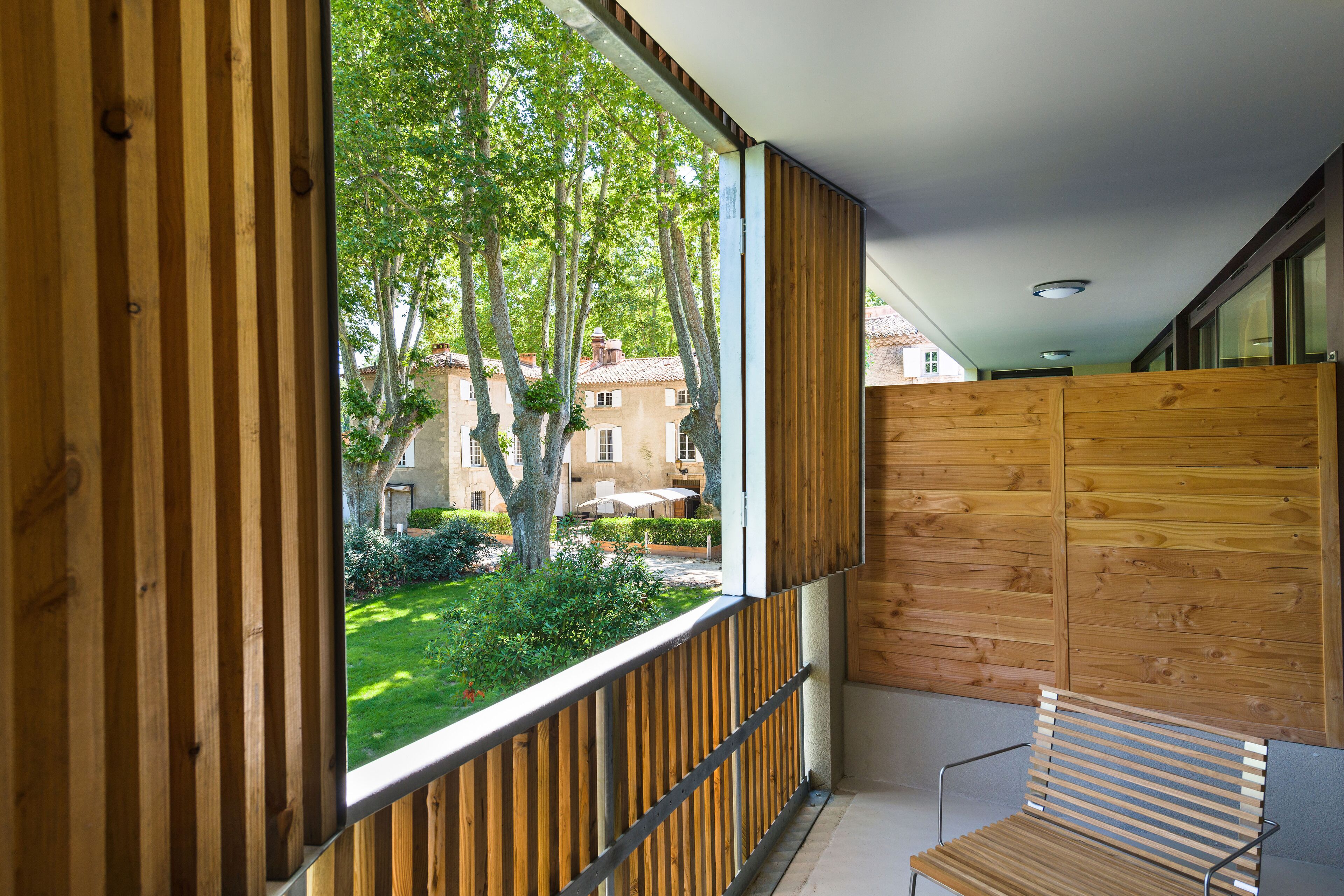 A balcony with wooden railings, a bench, and a view of a courtyard with trees and buildings.