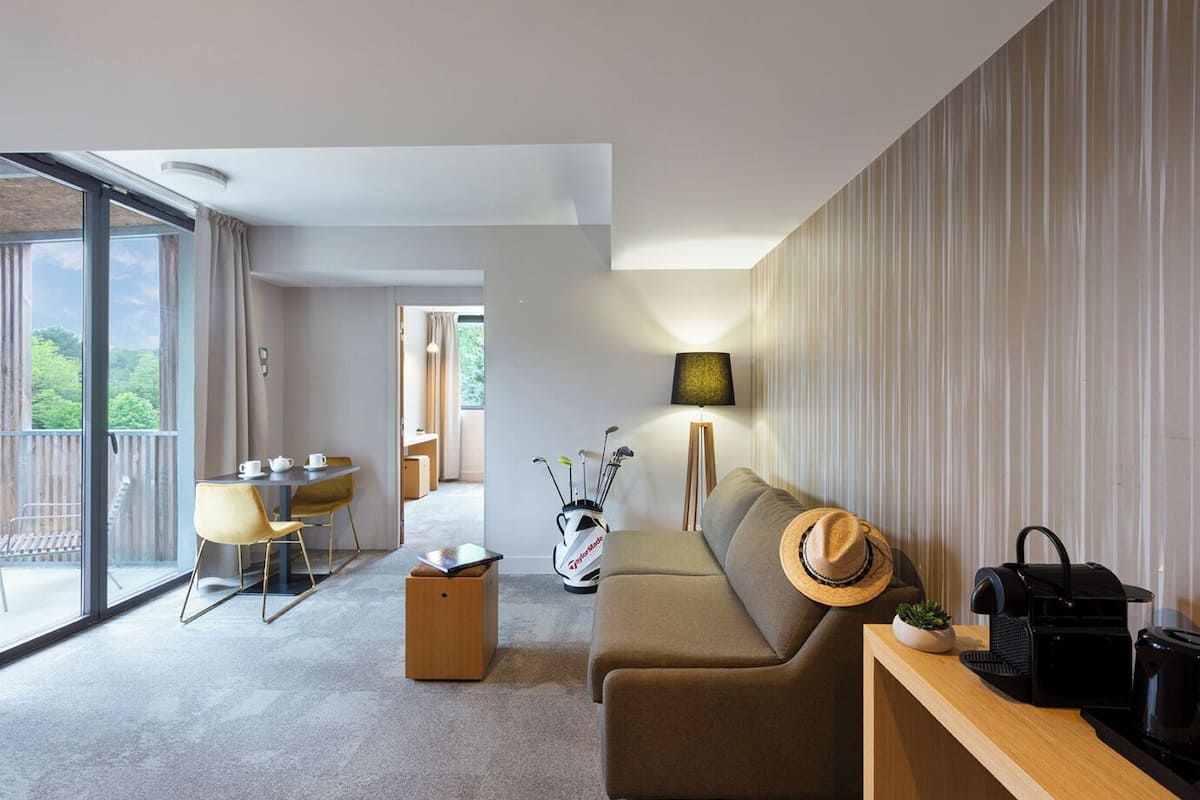 A modern living room with a sofa, a coffee table, a dining area with a table and chairs, a golf bag, and a view of greenery through large windows.