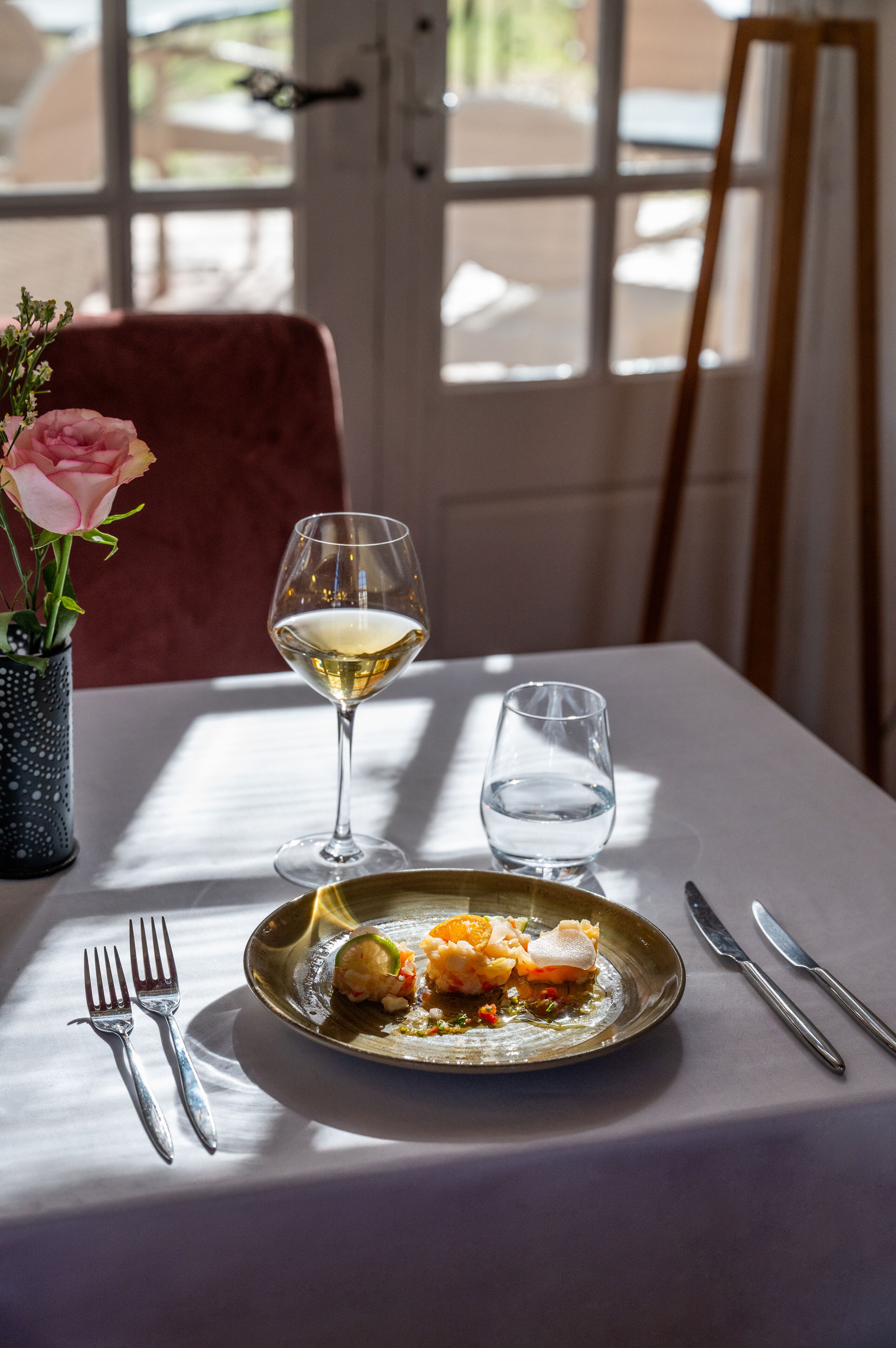 Una mesa de comedor con un plato de comida, una copa de vino blanco y un vaso de agua. Hay un florero con una flor rosa sobre la mesa.