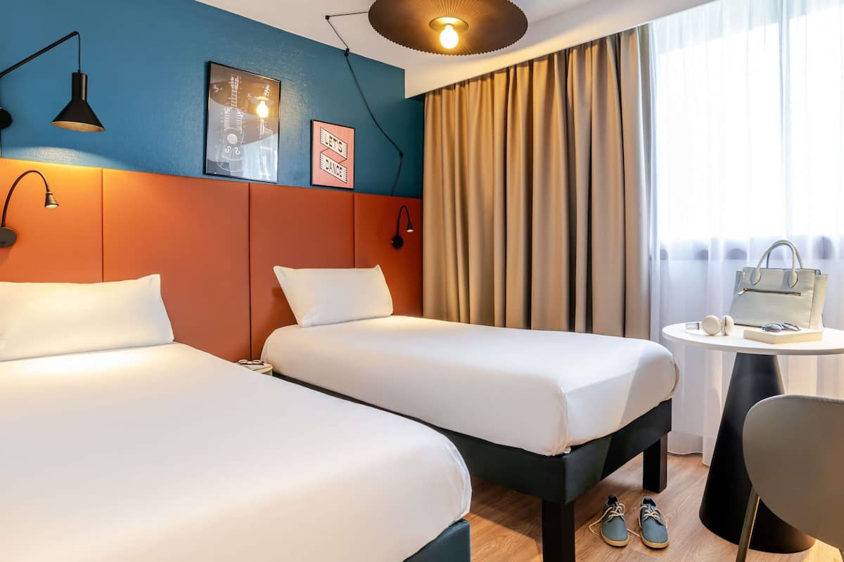 A hotel room with two beds, a desk, a chair, a lamp, a window with curtains, and a wall with framed pictures.
