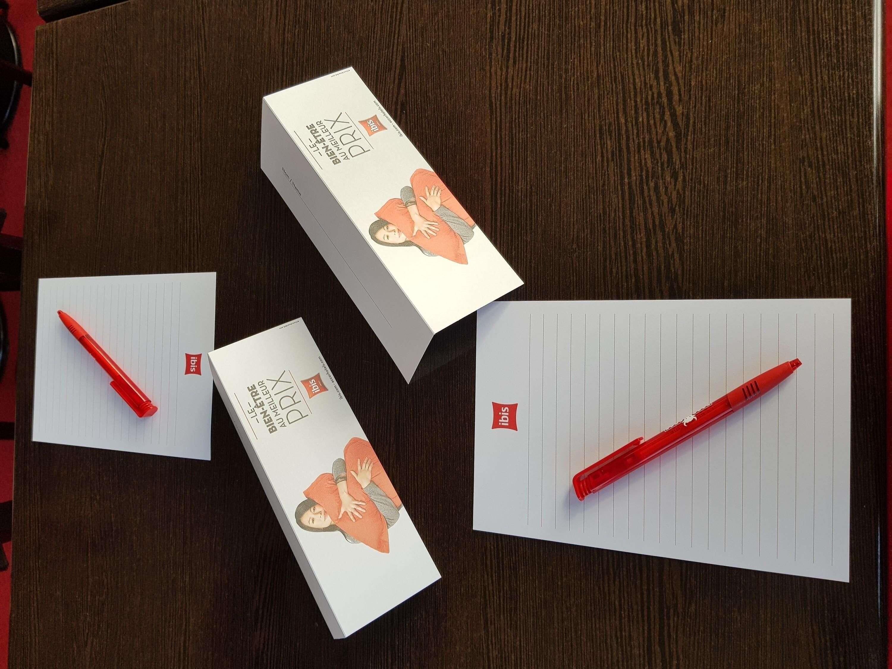 A box of red pens, a lined paper, and a notebook on a wooden surface.