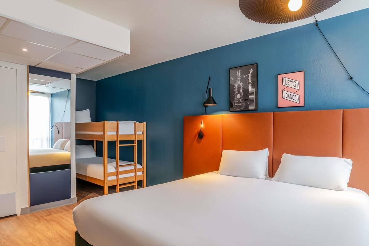A hotel room with a bunk bed, a large bed, a wall-mounted lamp, and two framed pictures.