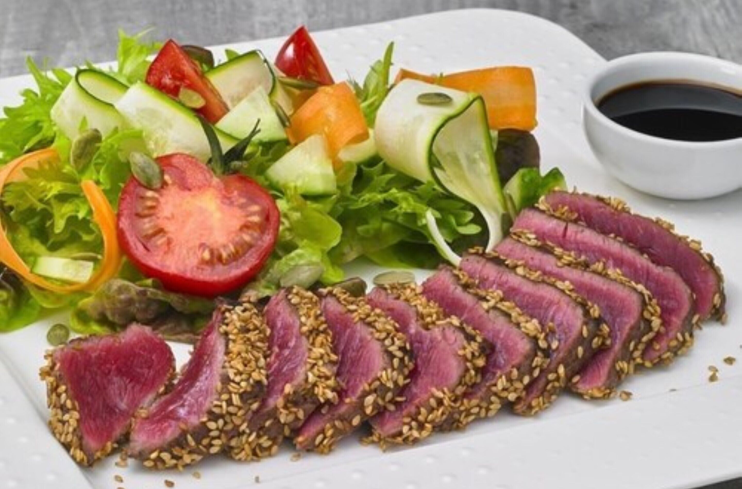A plate with sliced meat topped with sesame seeds, a mixed vegetable salad, and a small bowl of sauce.
