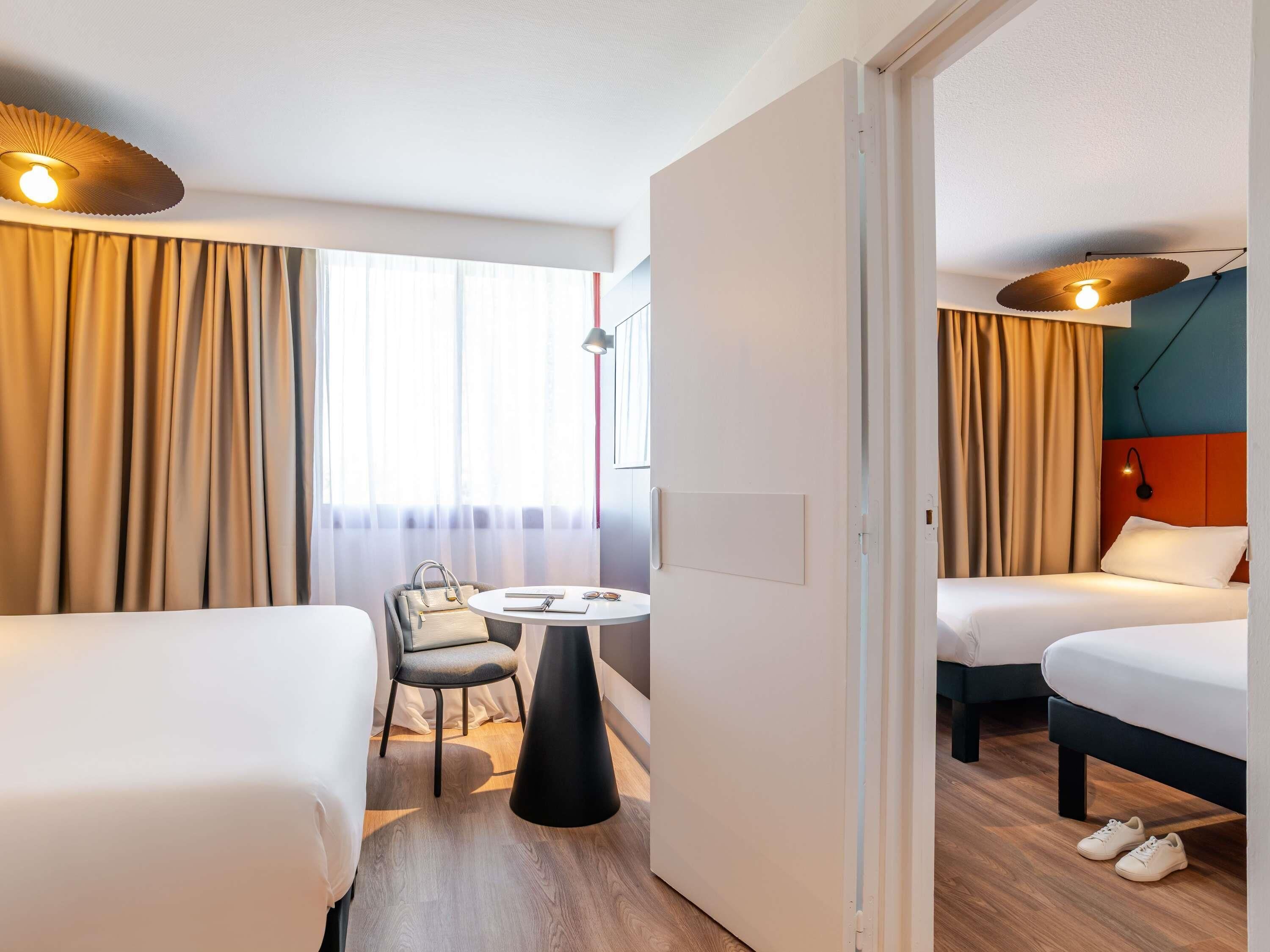 A hotel room with two beds, a small table with a chair, and a view of the outdoors through large windows.