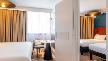 A hotel room with two beds, a small table with a chair, and a view of the outdoors through large windows.