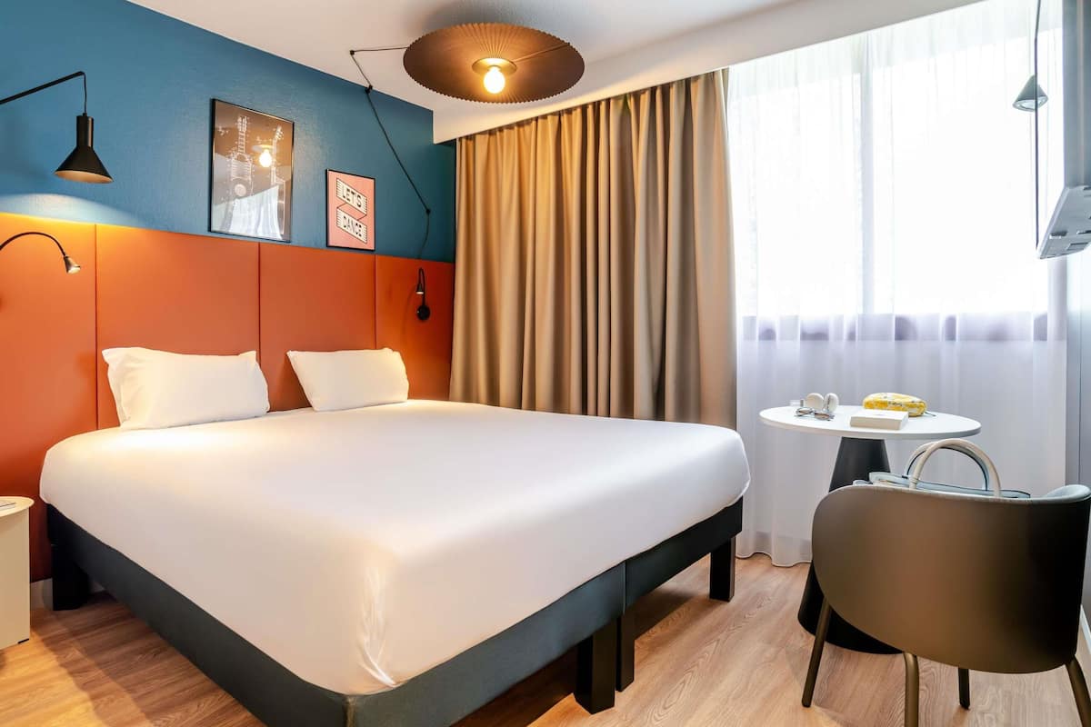 A hotel room with a large bed, a desk with a chair, a round table with a lamp, and a window with curtains.