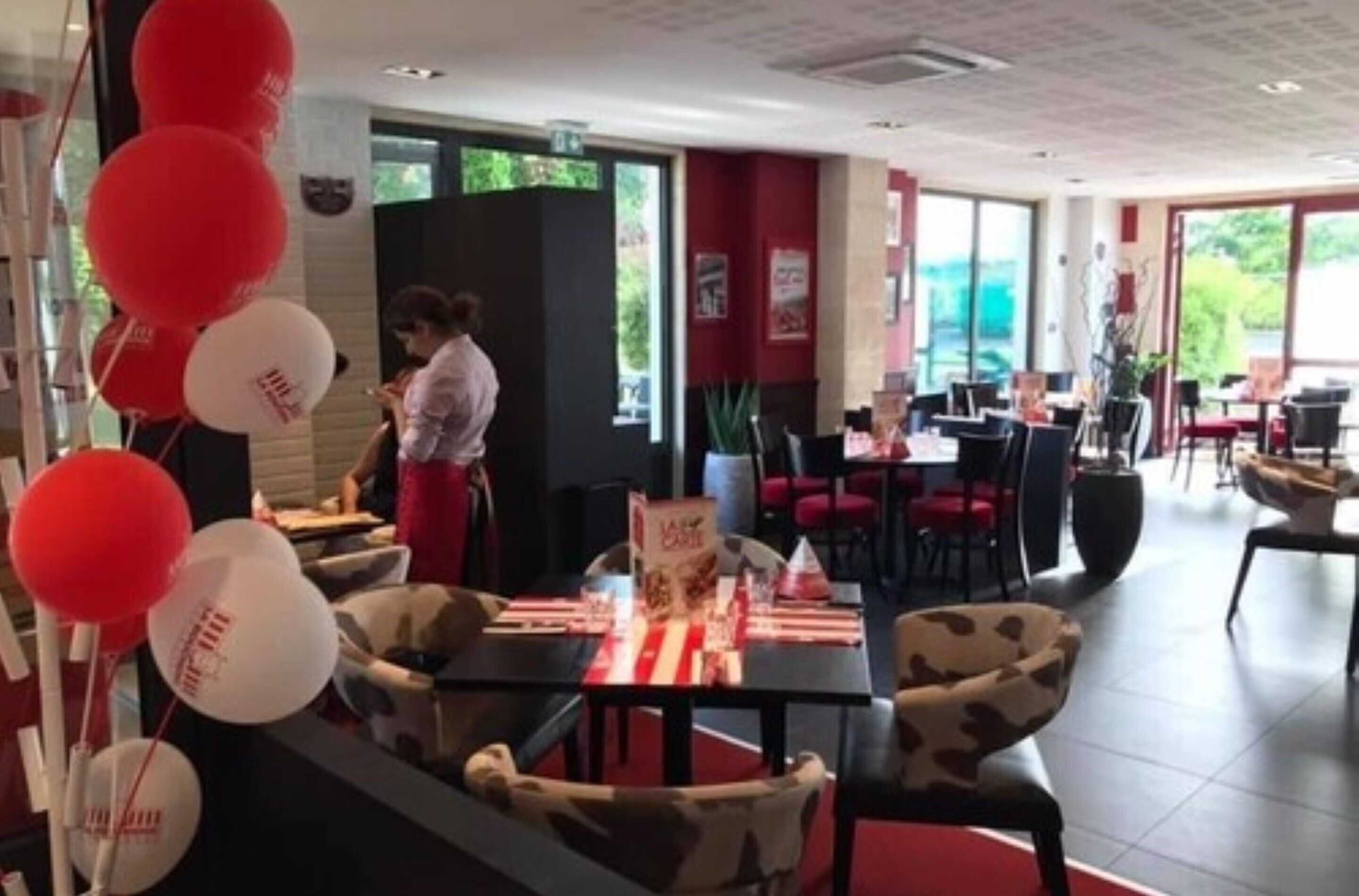 A restaurant with tables set for dining, a woman in the background, and red and white balloons.