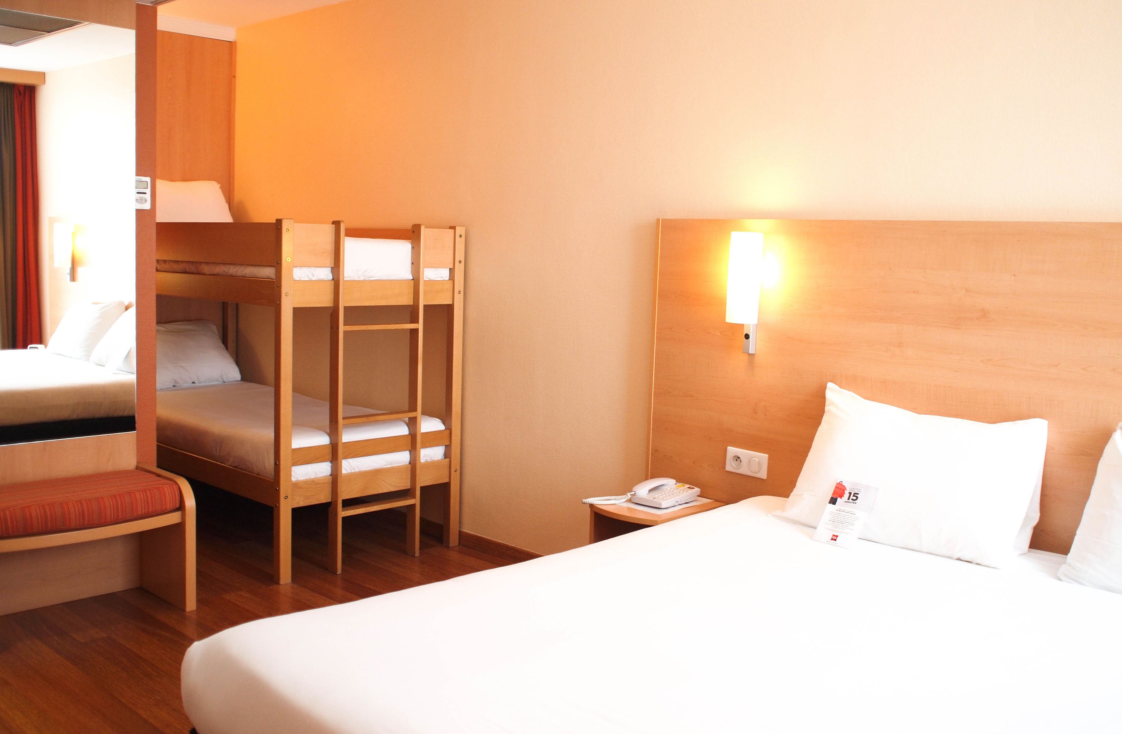 A hotel room with a bunk bed, a single bed, a bedside table with a phone, and a wall-mounted light.
