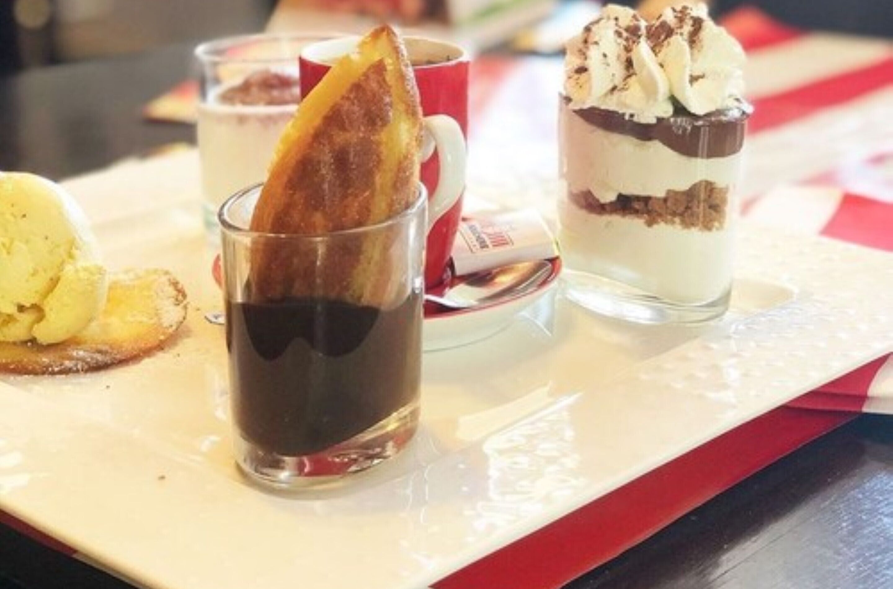 A dessert tray with various items including a glass of dark liquid, a slice of bread, and a layered dessert with whipped cream and chocolate.