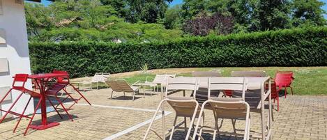 An outdoor seating area with tables and chairs, surrounded by a hedge and trees.
